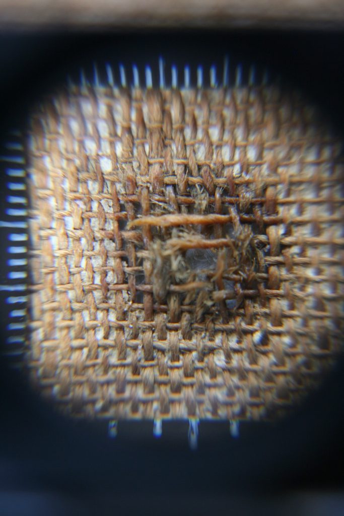 The interwoven threads of a canvas are seen through the lens of a microscope. A central area of damage is repaired with new threads of a slightly darker colour.