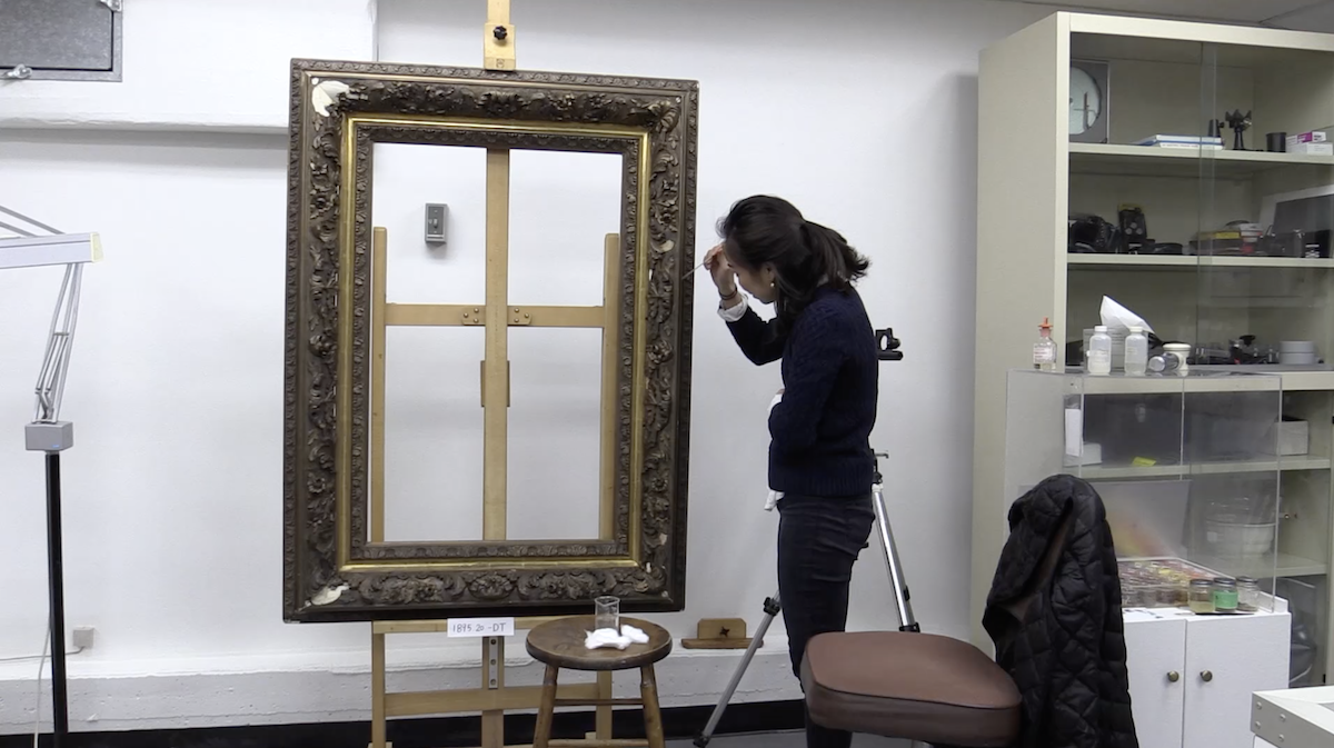 In a conservation laboratory, an adult stands next to a large frame, cleaning it with a tiny swab.