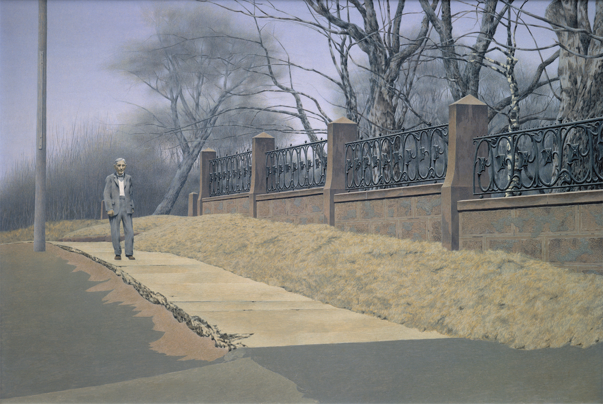 An older man with grey hair and a loose-fitting grey suit walks towards us on the sidewalk between the paved road and a decorative stone and wrought iron fence. Alongside him, the trees stand tall and bare, and the grass is dry and yellowed.