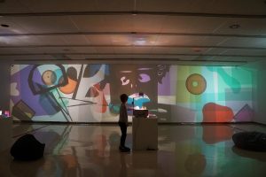 A child stands at an overhead projector, moving coloured shapes on its surface. In front of them, layers of colourful shapes are projected completely filling the gallery wall.