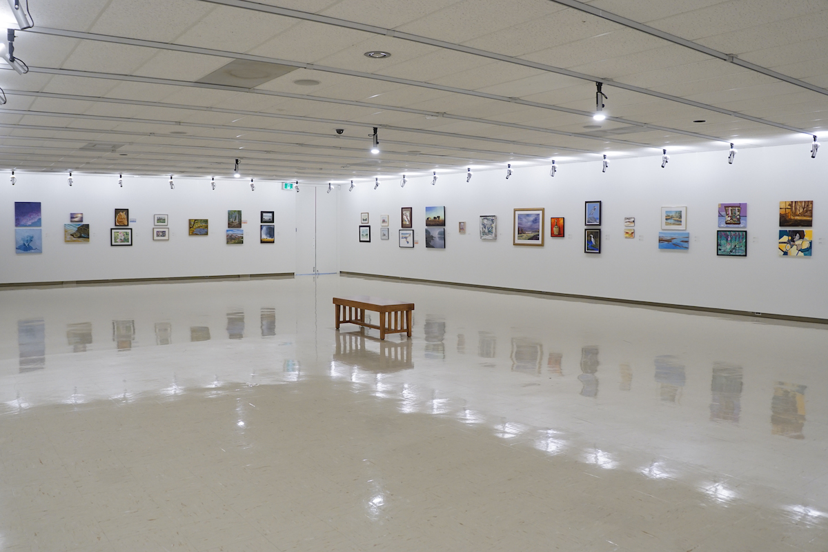 A large gallery with white walls display several artworks of varying sizes, media and content. In the centre of the room is a wooden bench.