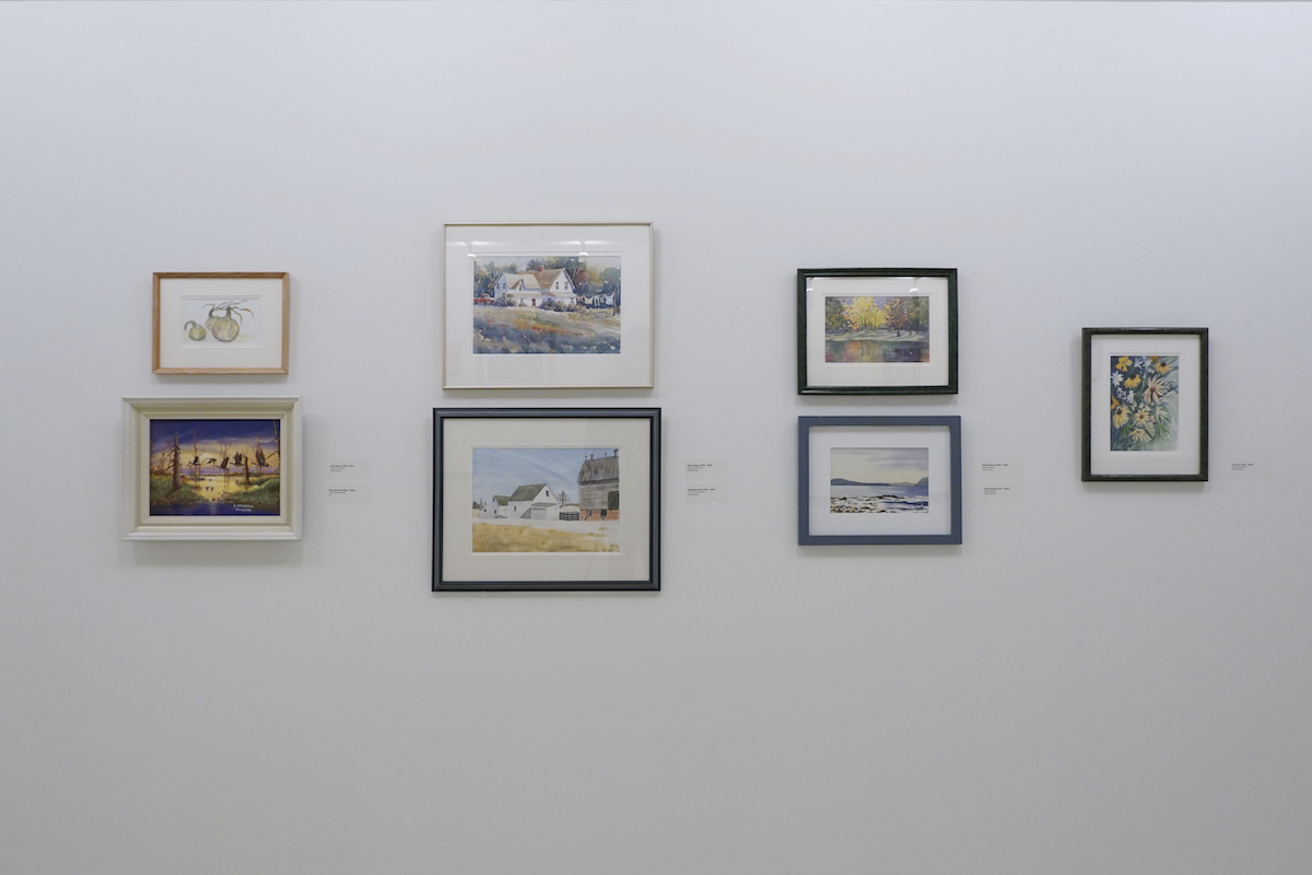 Seven framed paintings hang on a white gallery wall. Each is in a different coloured frame.