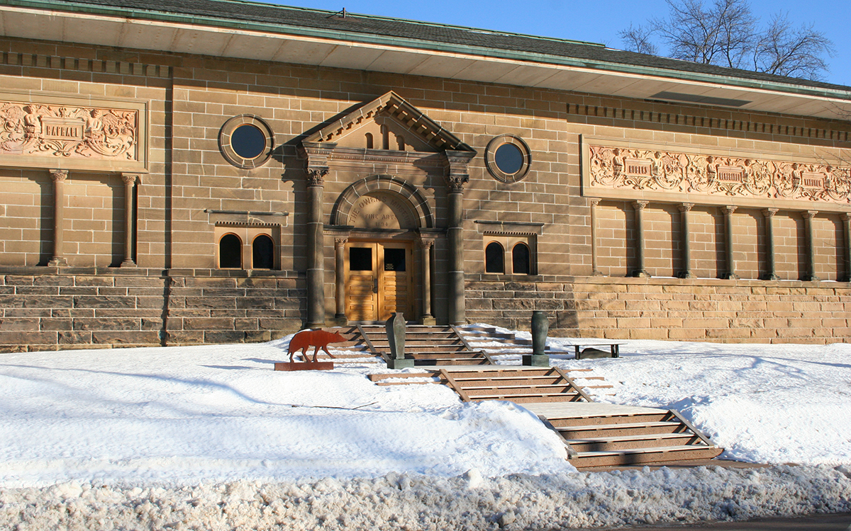 Snow surrounds a stone building with classical architectural features. On either side of the stairs leading to the door are sculptures of urns and a wolf.