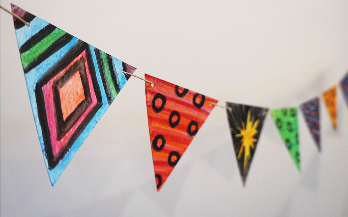 Triangles of painted paper are strung on twine. Each is painted with a different bright colourful pattern.
