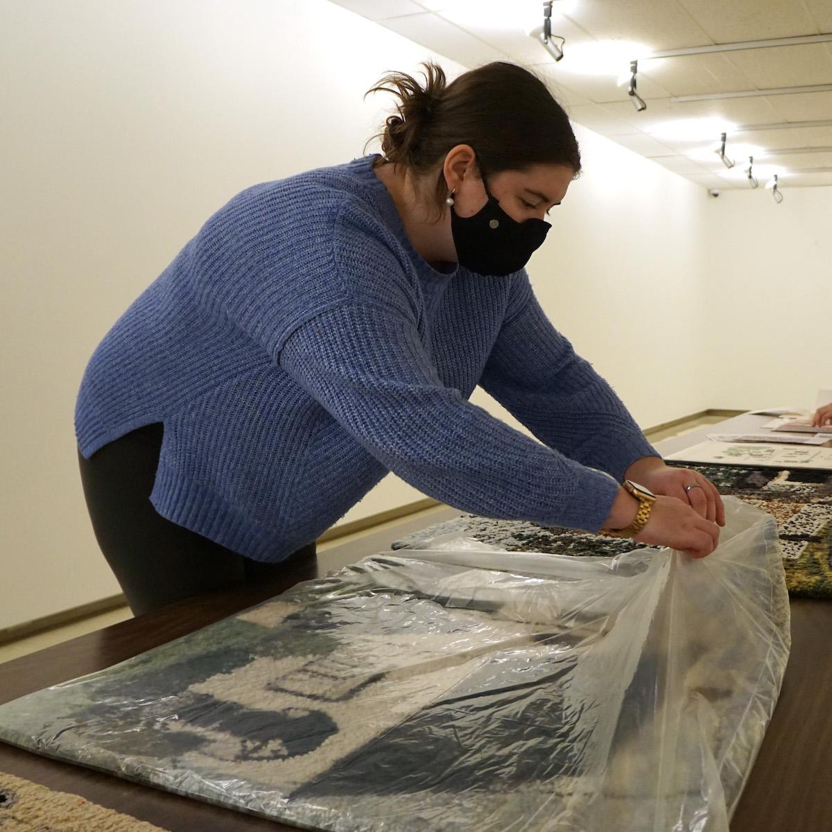 Paige leans over a table, unwrapping an artwork covered with thin plastic.