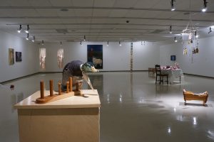 An exhibition featuring paintings, drawings, photographs, and sculptures is on display in a spacious gallery with white walls. In the foreground, a duck decoy with a bedazzled head on a wooden coat hanger rests on a plinth.