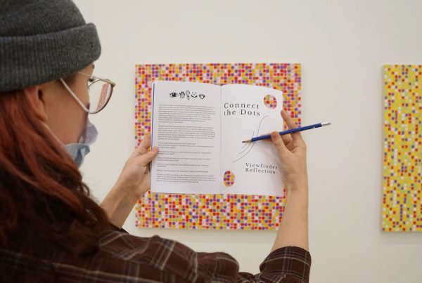 A student holds up a publication near a painting of a colourful grid on a gallery wall. The page on the left has several paragraphs of text. Two circles have been cut from the right page, allowing details of the painting to peek through.