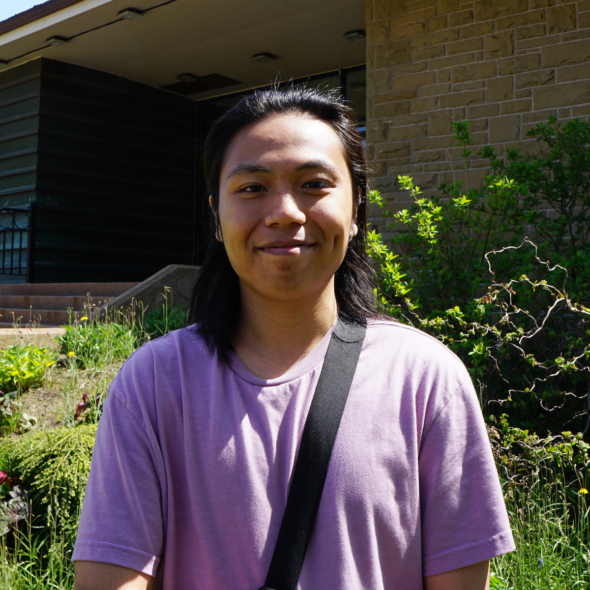 Ranz stands smiling, wearing a pink T-shirt and cross body bag, in front of the Owens. The garden behind him is thriving with leafy green plants and small yellow flowers.
