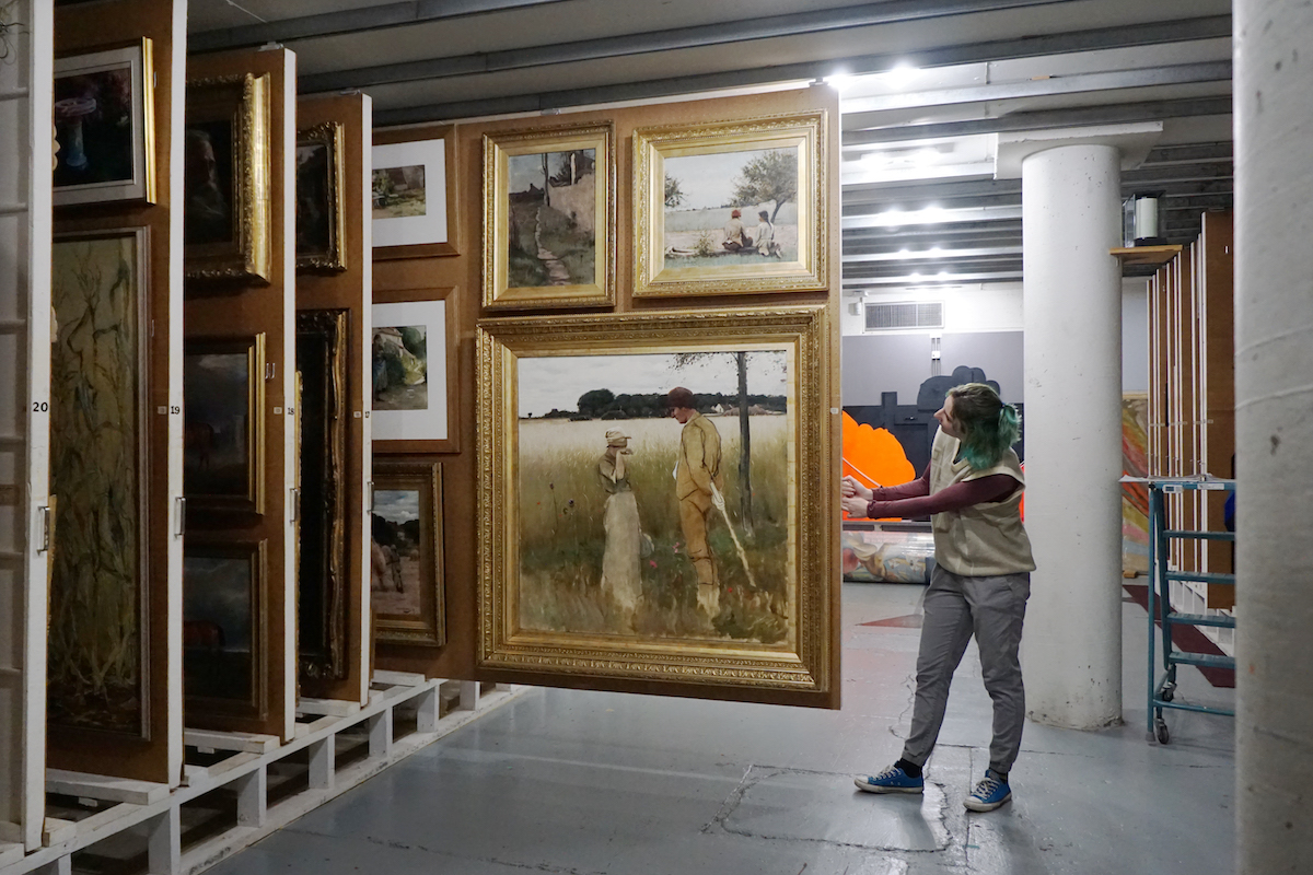 In a large painting vault, an adult pulls a panel revealing several paintings in gold frames.