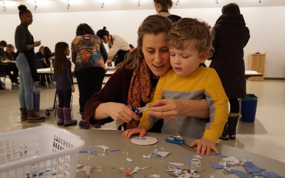 In a gallery an adult and child sit together at a table cluttered with small scraps of cut paper. The adult uses a pair of scissors to cut a small pieces of paper while the child watches. Behind them adults and children work at other tables.