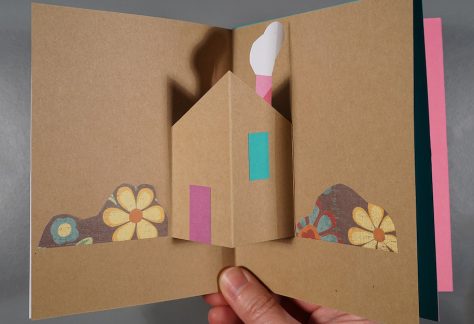 A hand holds a pop-up book. A house pops out from the brown pages, decorated with floral patterned bushes, and a pink chimney and front door.