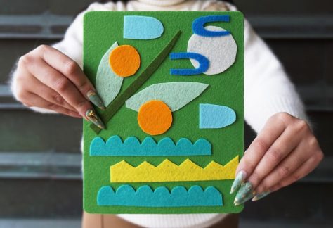 Two hands hold up a colourful artwork made of fuzzy felt. On the green background, cut shapes make a sky with moon and clouds and a tree branch with oranges above three cut silhouettes of waves.