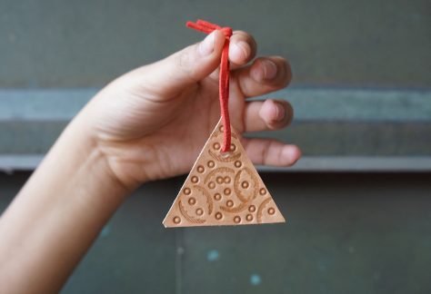 A hand holds up a small leather ornament, dangling from a red suede string. The triangular leather shape is stamped with small circles and larger striated curved forms.
