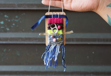 A hand holds up a small woven tapestry on a wooden backing. The weaving of colourful yarn and ribbon is adorned with beads and a pop can pull tab. Blue tassels dangle from the bottom.