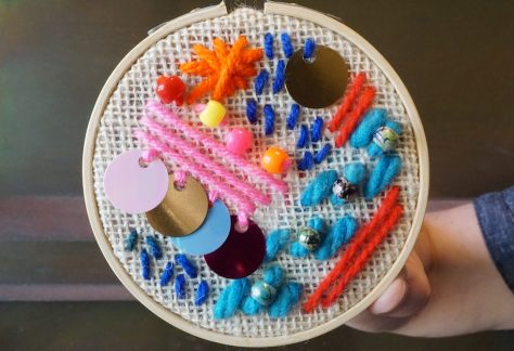 An embroidery hoop is filled with colourful yarn, beads, and sequins on beige burlap fabric. A hand holds the hoop in front of a copper wall.