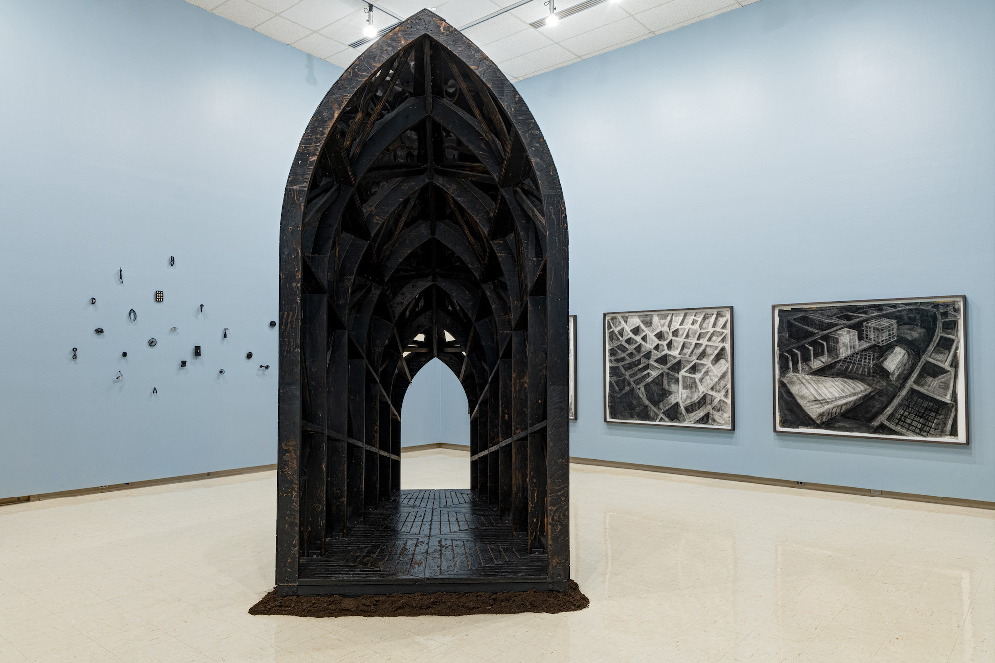 Looking into a gallery with high, blue walls, a large wooden, cathedral shaped sculpture fills the space. On the walls behind are several small sculptures, and three large, geometric drawings.