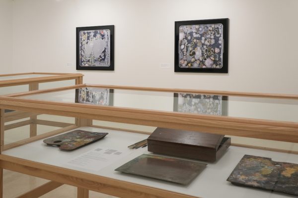 In a gallery, two wooden display cases hold wooden boxes and palettes. On a wall behind the case, are two framed artworks of petri-dishes with bacteria colonies.