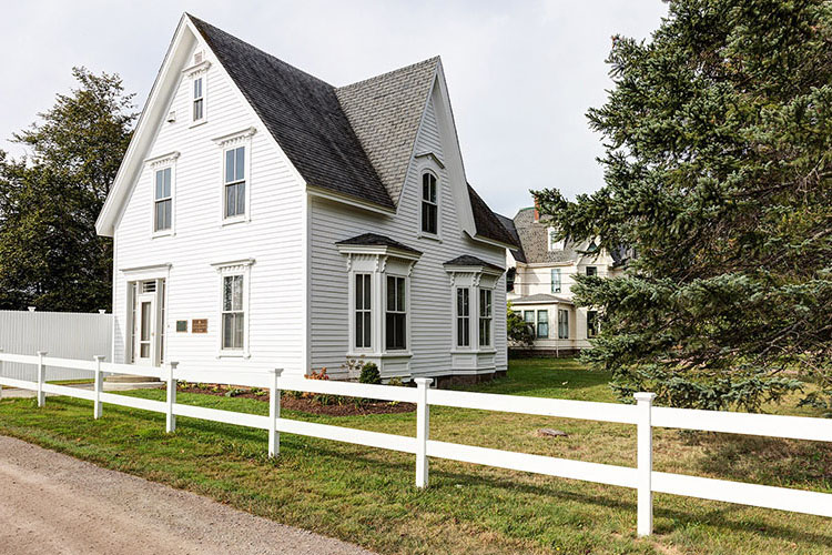 A low white fence runs parallel to a driveway that leads to a two-storey Carpenter Gothic-style, white clapboard house with tall windows, a steeply pitched roof and a prominent cross-gable.