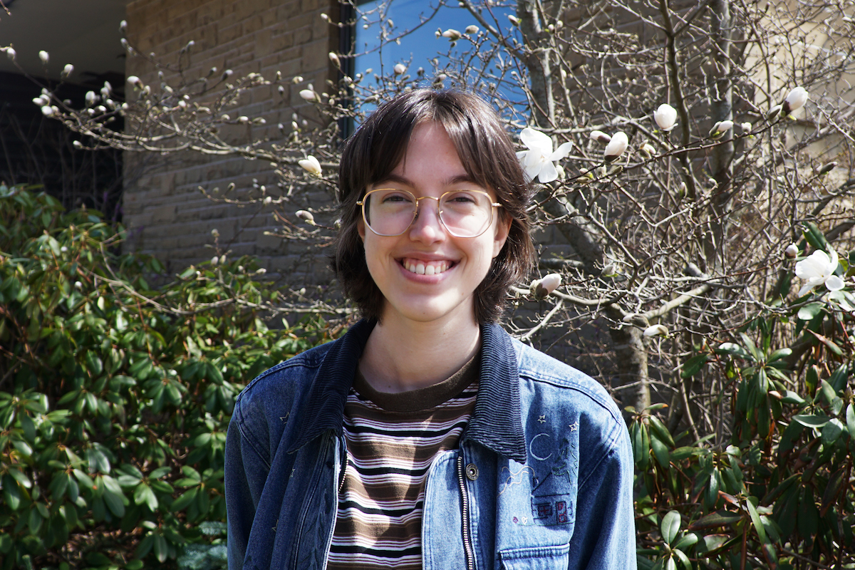 Caitlin Gallant stands in front of a flowering bush, facing the camera and smiling. They have short brown hair and glasses.