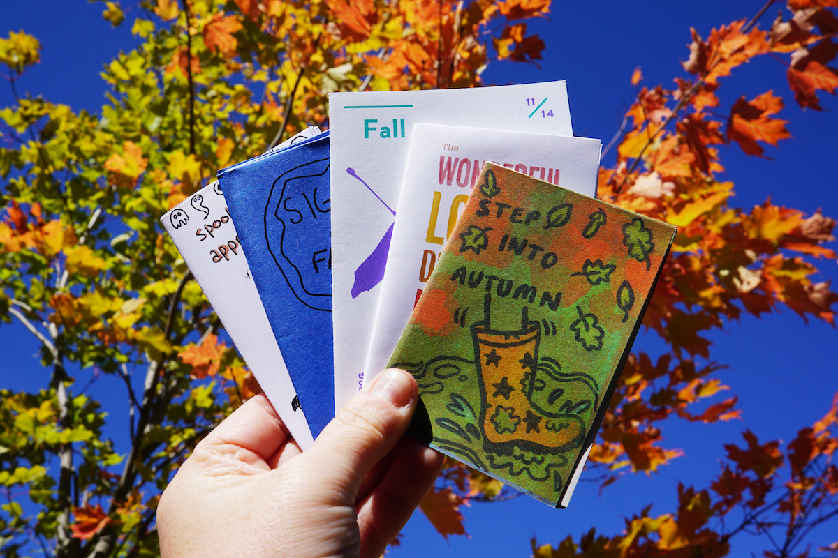 A hand holds five colourful zines in front of a maple tree with red, orange, yellow and green leaves. The frontmost zine features falling leaves, and a boot splashing in a puddle with the text "step into Autumn".