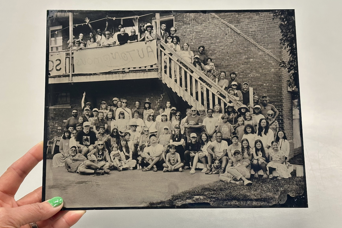 A hand holds a black and white group photograph showing a large gathering of people posed in front of and on an outdoor stairway attached to a brick building.