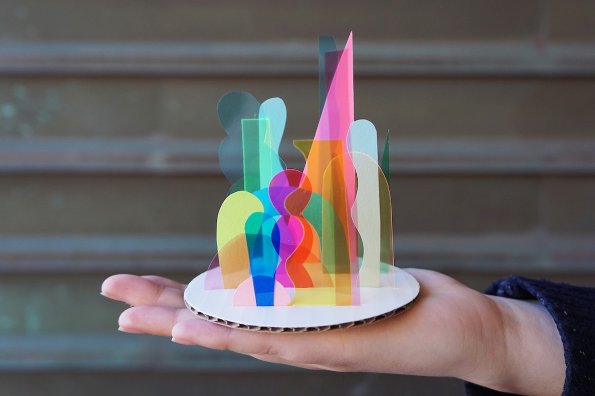 A hand holds a round cardboard platform with layered, multicoloured translucent abstract shapes.